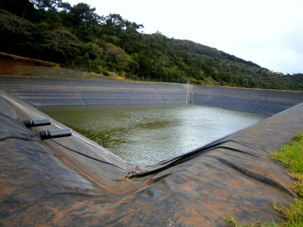 A lined artificial pond with two black pipes, surrounded by grassy and wooded hills.
