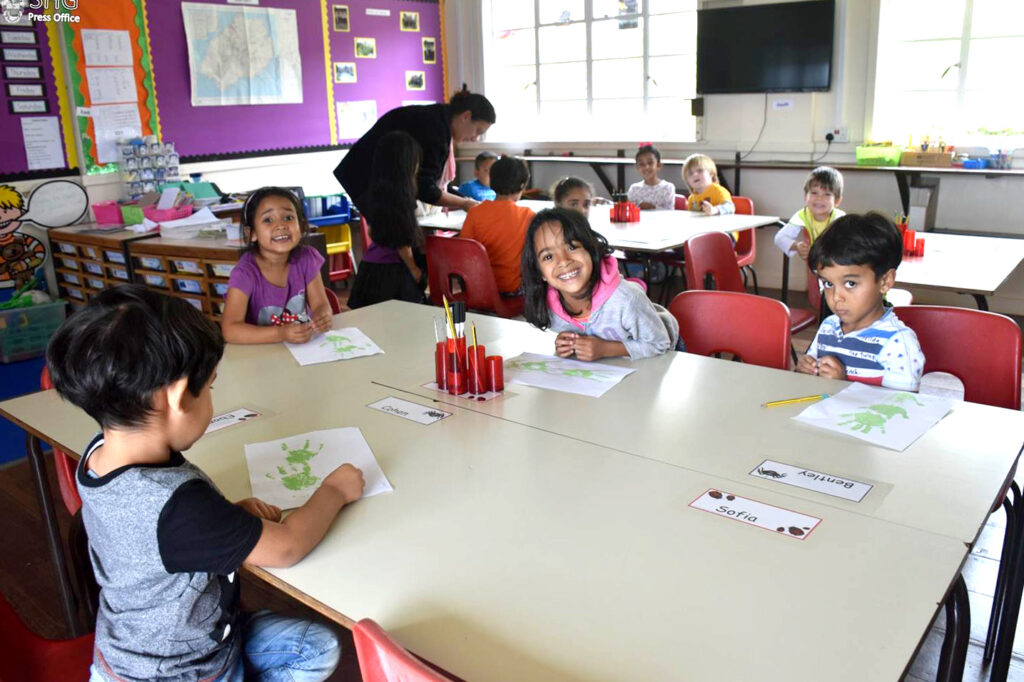 Young children sitting at classroom tables, drawing and smiling, with a teacher assisting in the background.