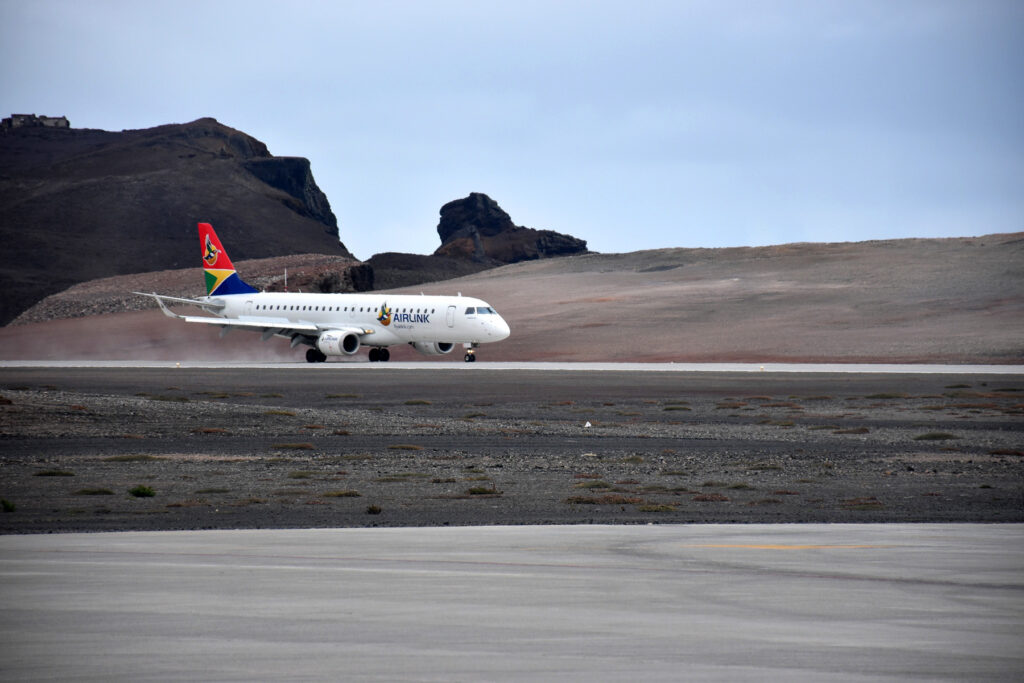 A white aeroplane with a colourful tail lands on a remote, rocky runway with hills in the background.