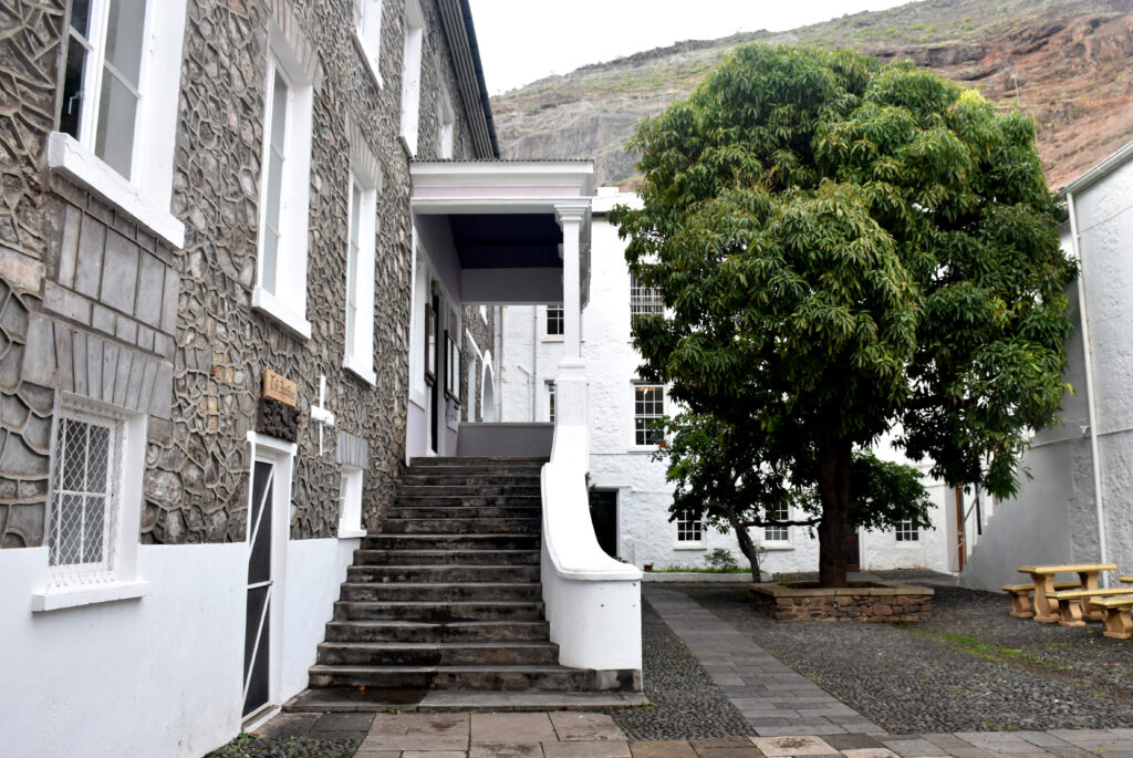 Castle with steps, a large tree, and picnic benches in a courtyard with a rocky hill in the background.