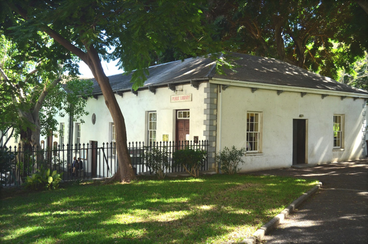 A white, one-storey library building with large windows and trees casting shade over the front lawn.