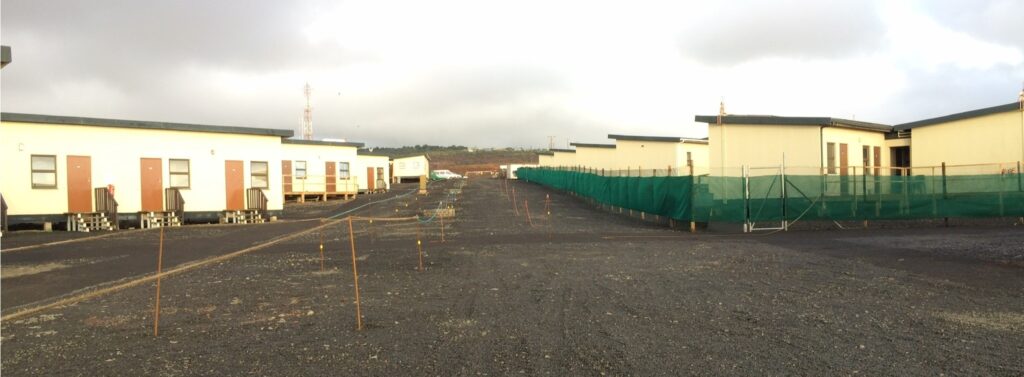 A row of temporary buildings with green fencing and an empty gravel path under cloudy skies.