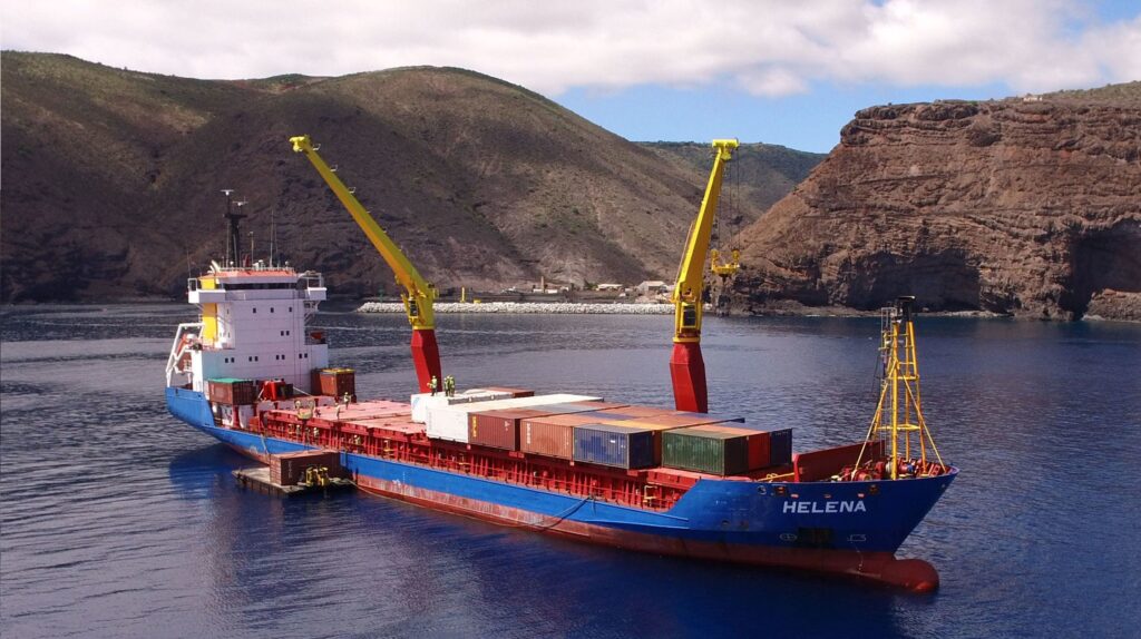 A cargo ship named HELENA with containers docks near rocky, green hills under a partly cloudy sky.