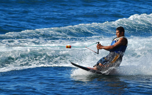 A man in a life jacket water skiing on blue water, holding onto a tow rope with waves behind him.
