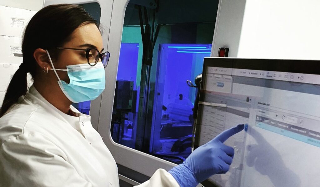 A scientist in a mask and gloves uses a touchscreen in a laboratory with medical equipment in the background.