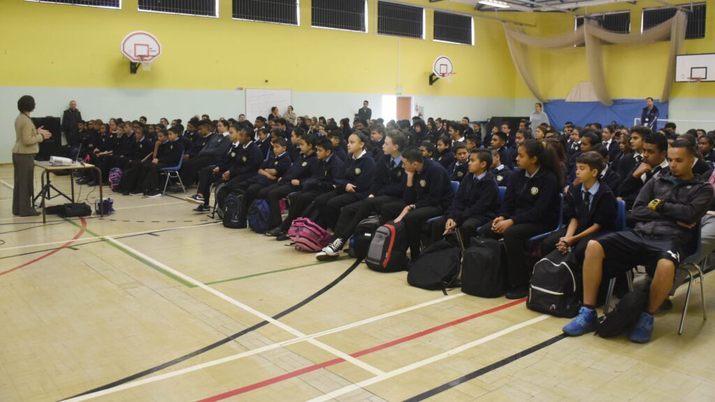 Students in school uniforms sit in a gymnasium, listening to a speaker at the front of the room.