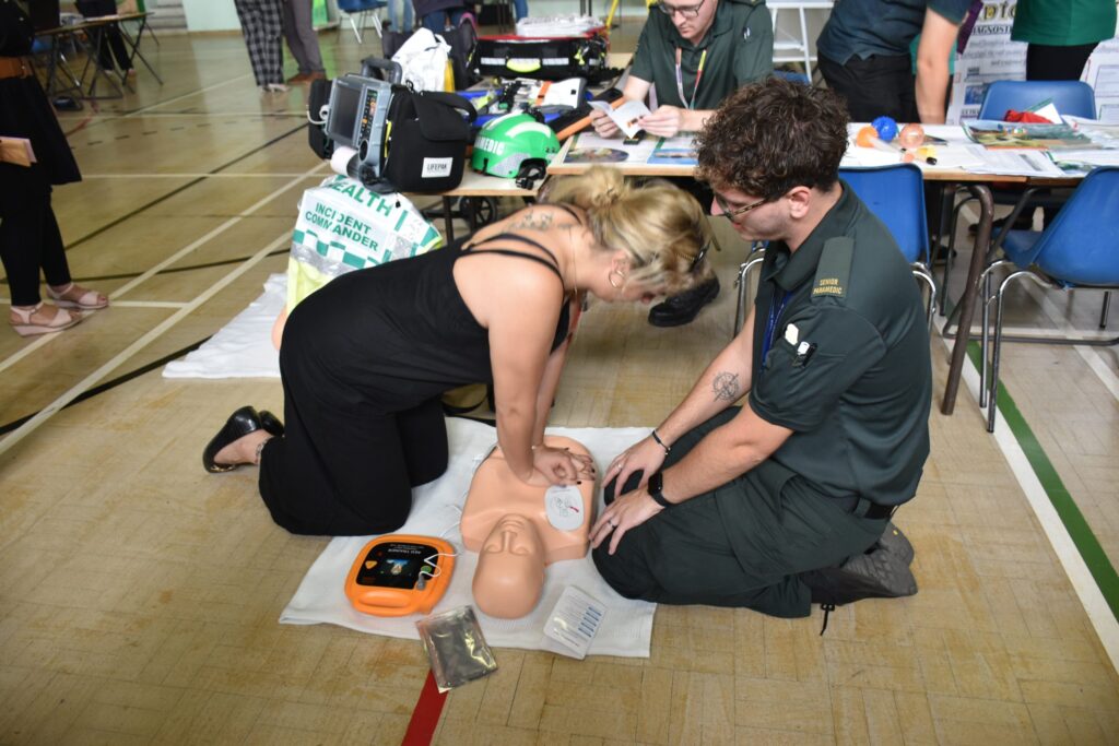 A woman practises CPR on a dummy with guidance from a paramedic in a classroom setting.