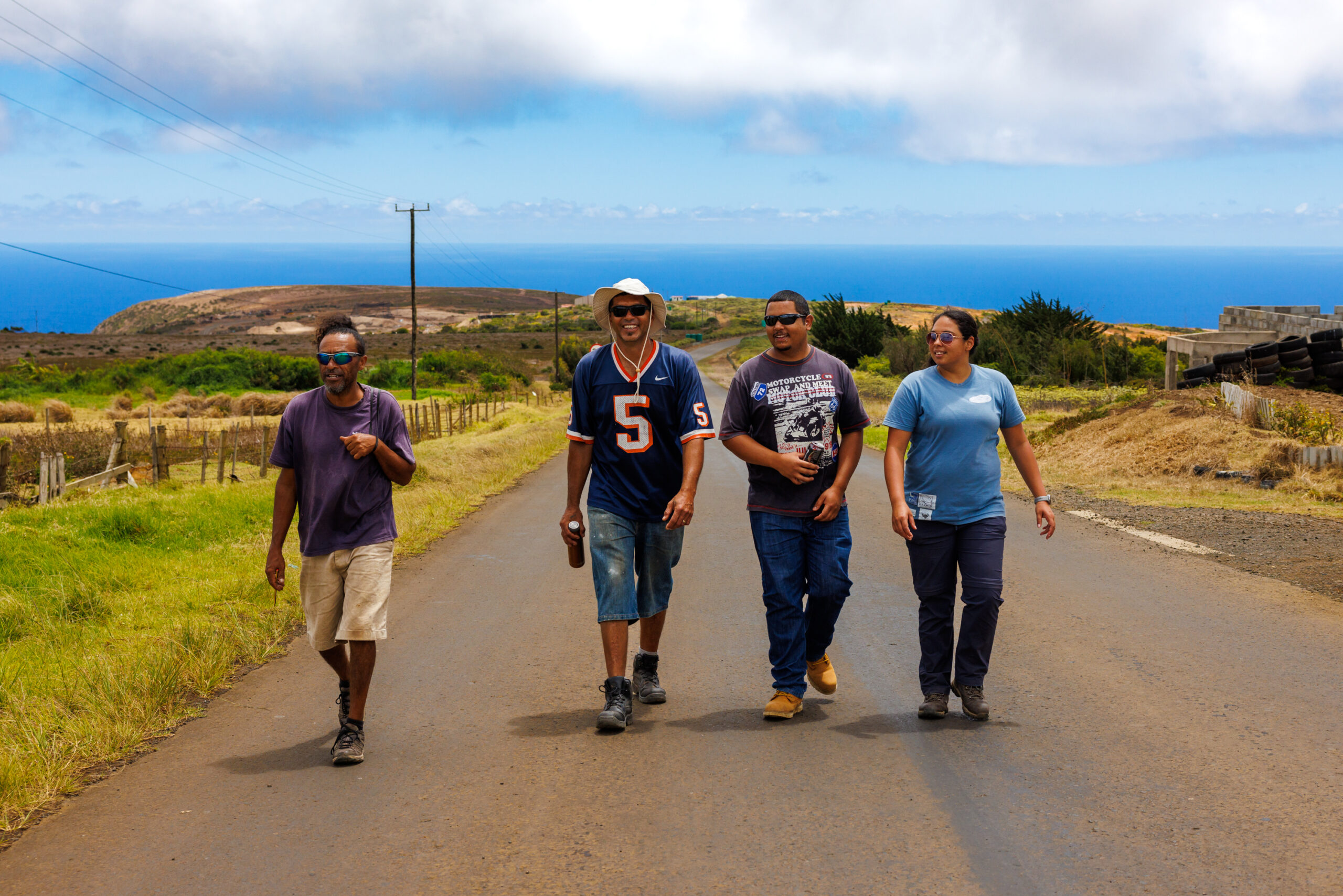 Four people walk together on a rural road with fields and sea in the background under a partly cloudy sky.