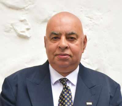 Bald man in a dark suit and patterned tie standing against a white textured wall, looking at the camera.
